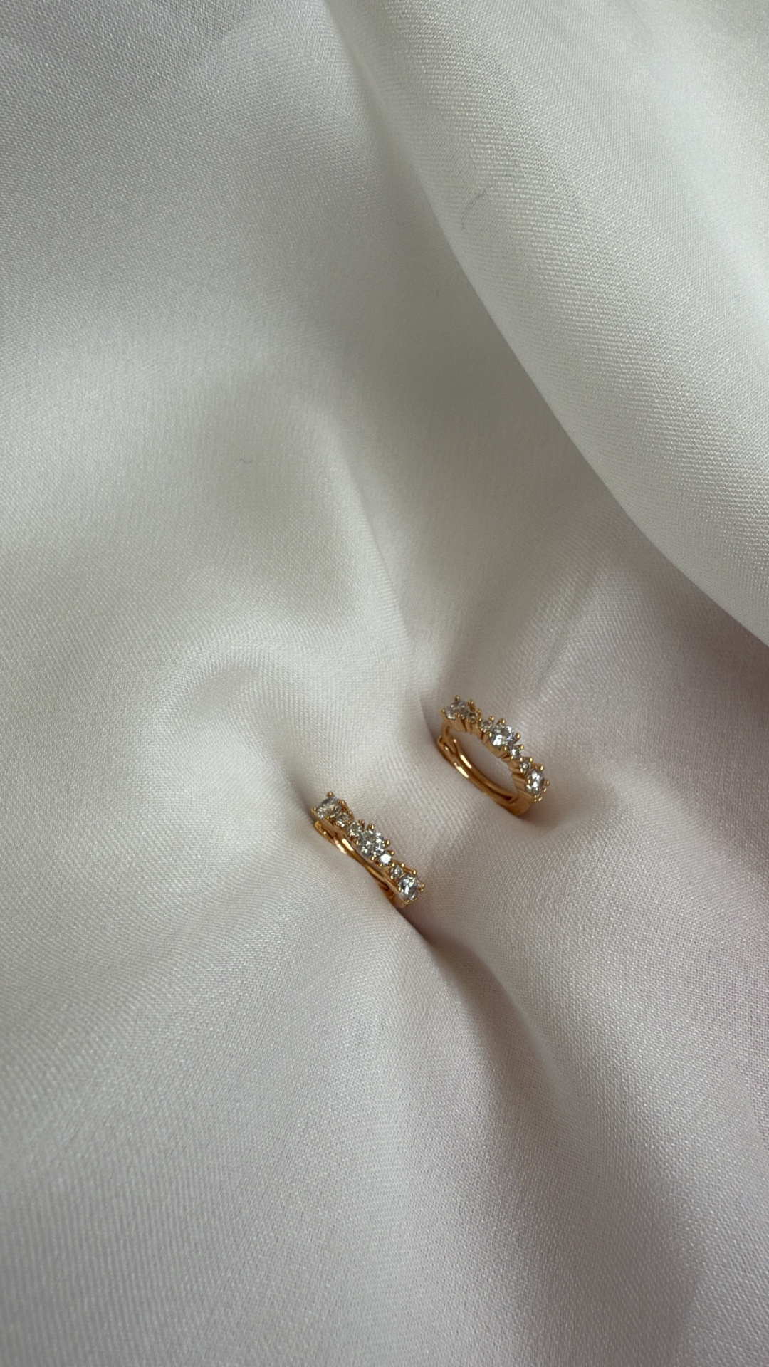 Brincos de argola dourados com pedras transparentes sobre tecido branco.