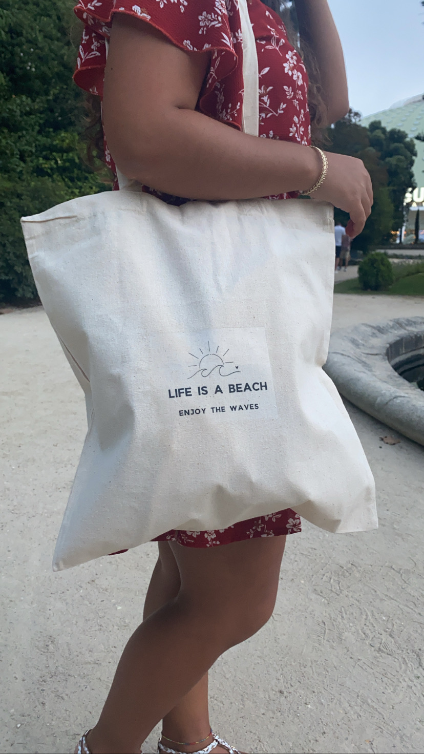 Bolsa de tecido creme com texto 'LIFE IS A BEACH ENJOY THE WAVES' e desenho de sol e ondas, usada por pessoa com vestido vermelho de flores brancas.