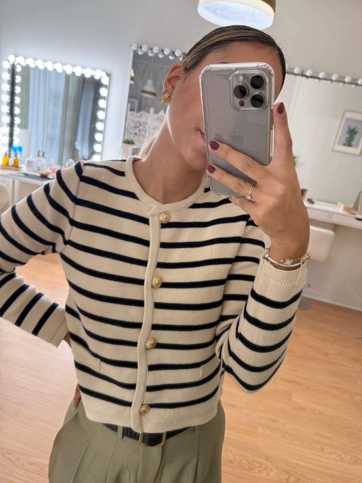 Cardigan creme com riscas pretas e botões dourados, calças verdes, ambiente interior com espelho iluminado