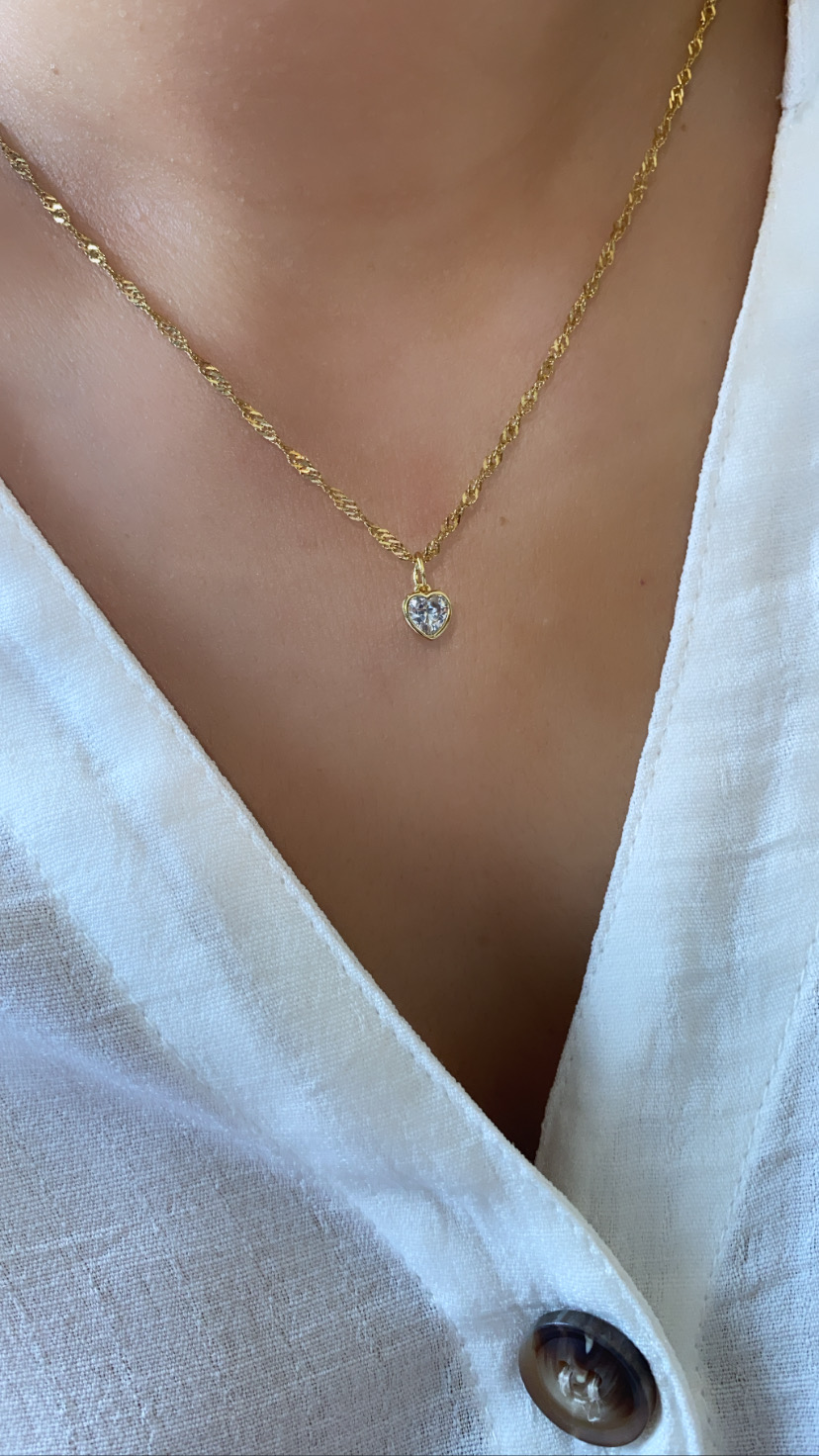 Colar dourado com pendente de coração de pedra transparente num pescoço com camisa branca