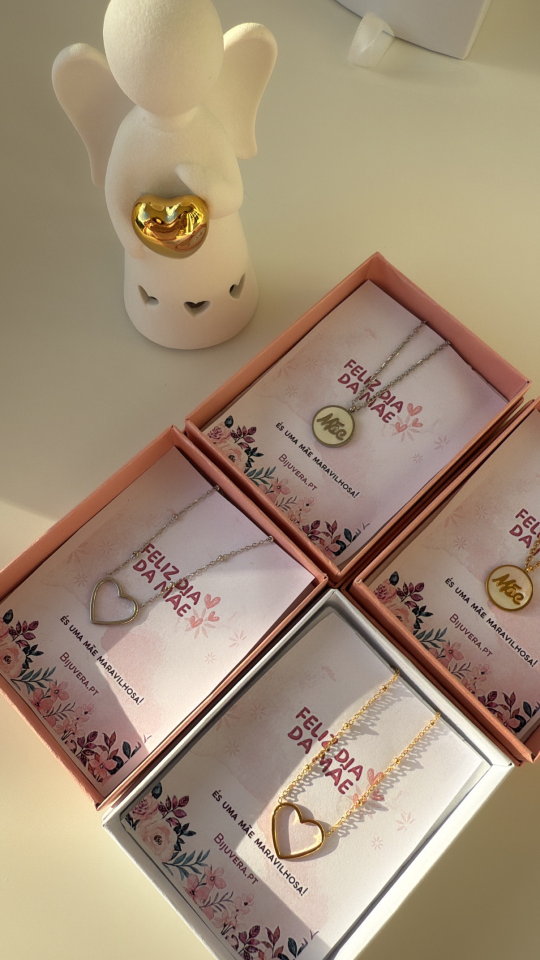 Colares em caixas cor-de-rosa com cartão Feliz Dia da Mãe e anjo decorativo branco com coração dourado.