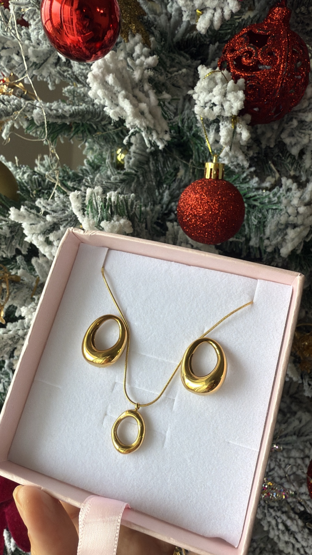 Conjunto de joalharia dourado com colar e brincos em forma oval numa caixa e árvore de Natal ao fundo