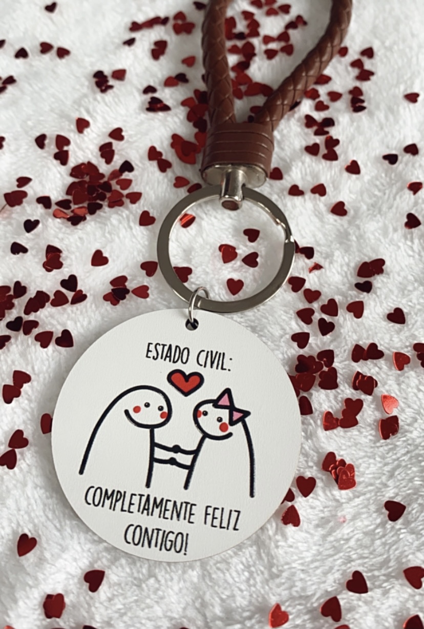 Chaveiro redondo branco com desenho de casal e texto em português sobre superfície branca com confetes vermelhos.