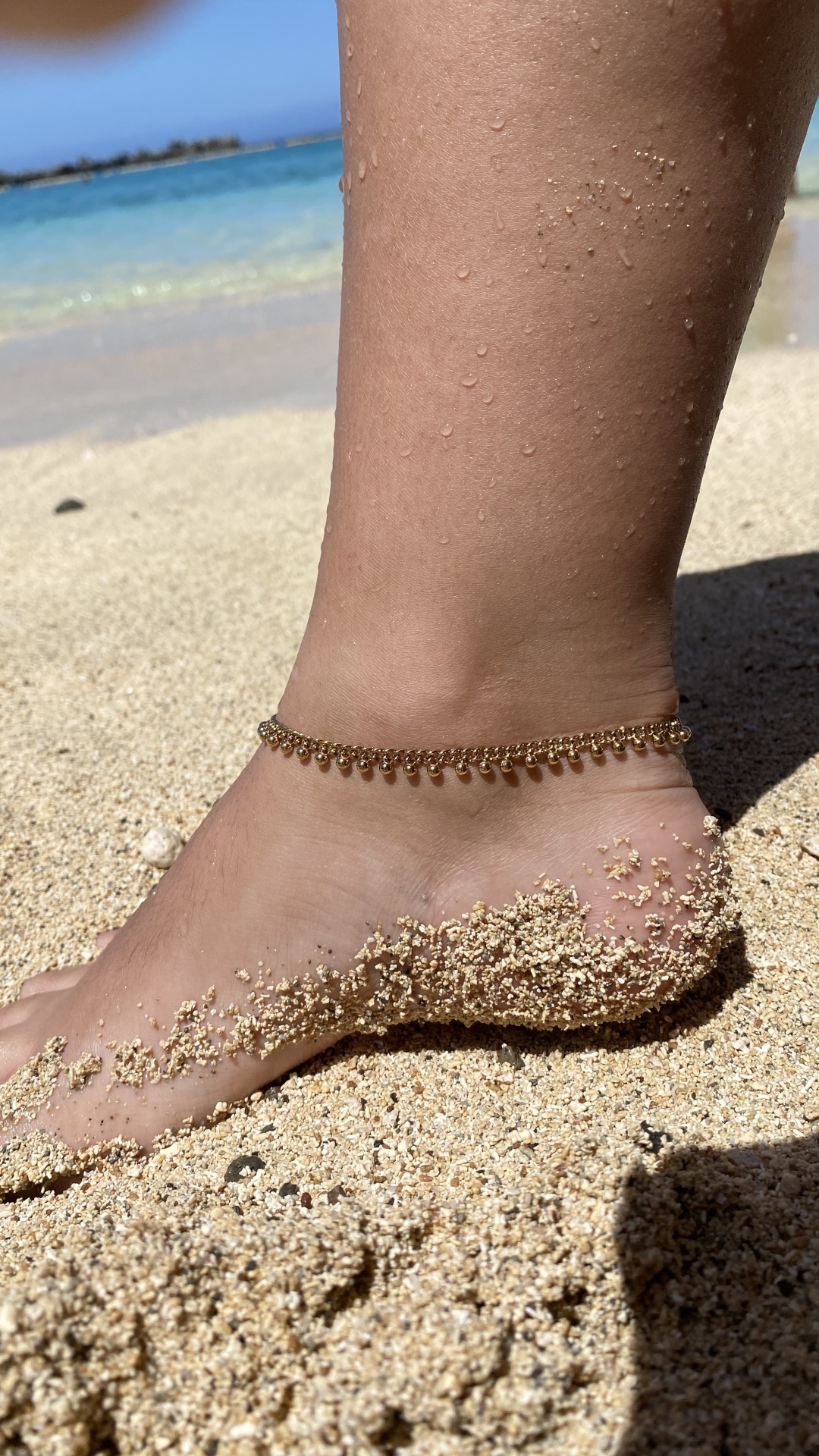Tornozelo com pulseira dourada em areia de praia junto ao mar