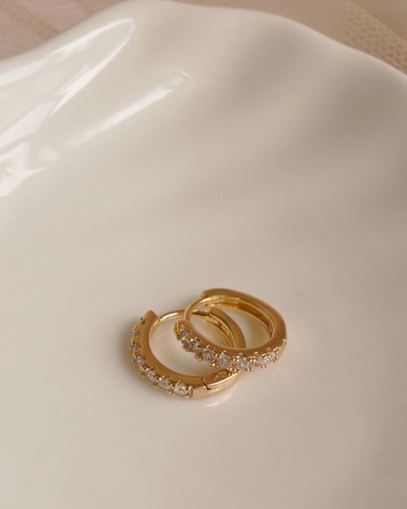 Brincos de argola dourados com pedras brancas numa superfície branca lisa