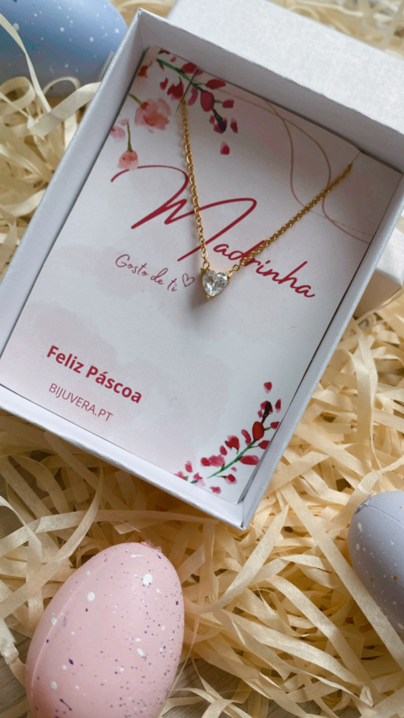 Colar dourado com pendente de coração numa caixa branca com cartão de Páscoa e decoração com ovos e palha.