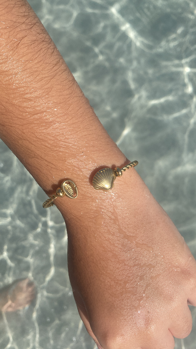 Pulseira dourada com pingentes de concha num pulso sobre água cristalina