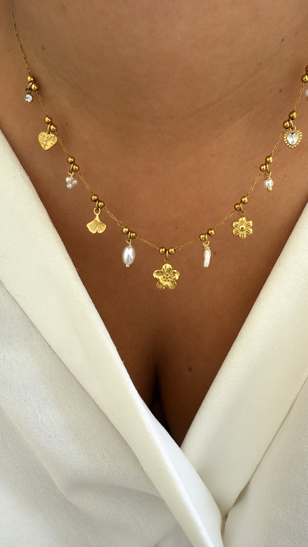Colar dourado com pingentes de flores, corações e pérolas, em pescoço com roupa branca