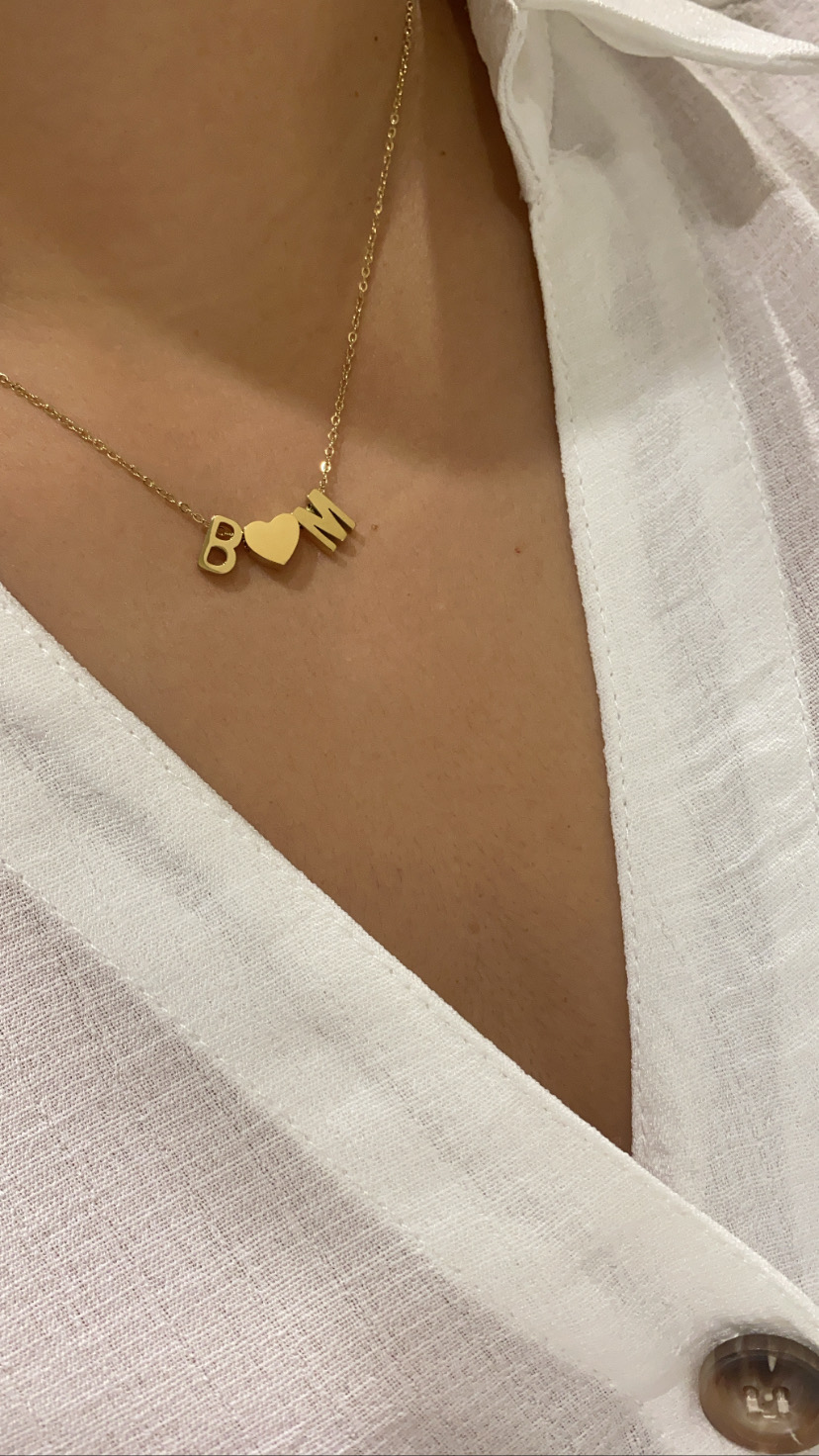 Colar dourado com letras B e M e coração entre elas, usado com camisa branca