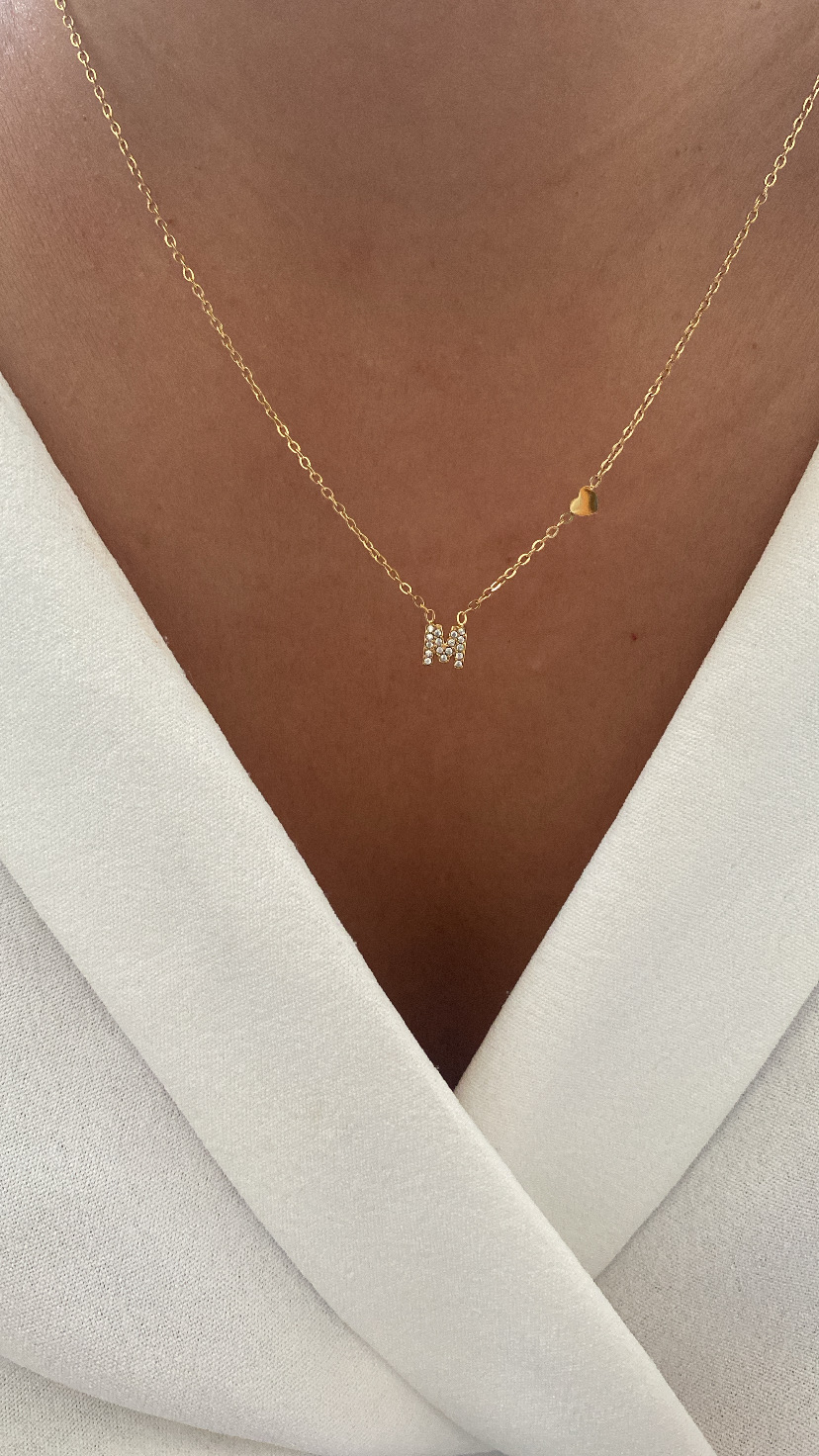 Colar dourado com letra M em cristais e coração, usado no pescoço sobre vestido branco