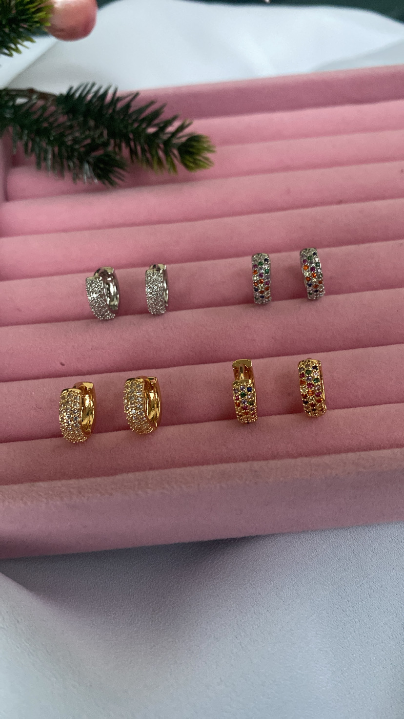 Oito pares de brincos em metal prateado e dourado com pedras brilhantes, organizados em suporte de veludo rosa.