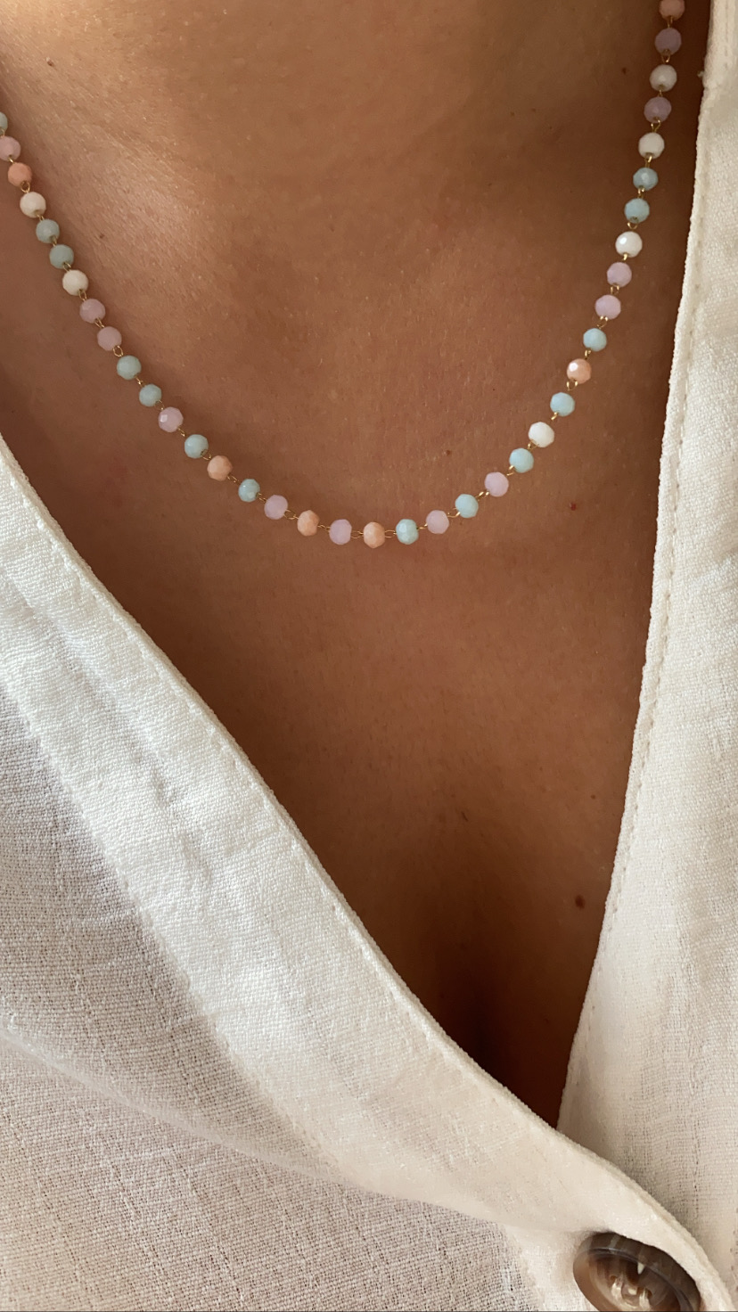 Colar com contas coloridas pastel e esferas douradas na pele, com camisa branca de tecido leve