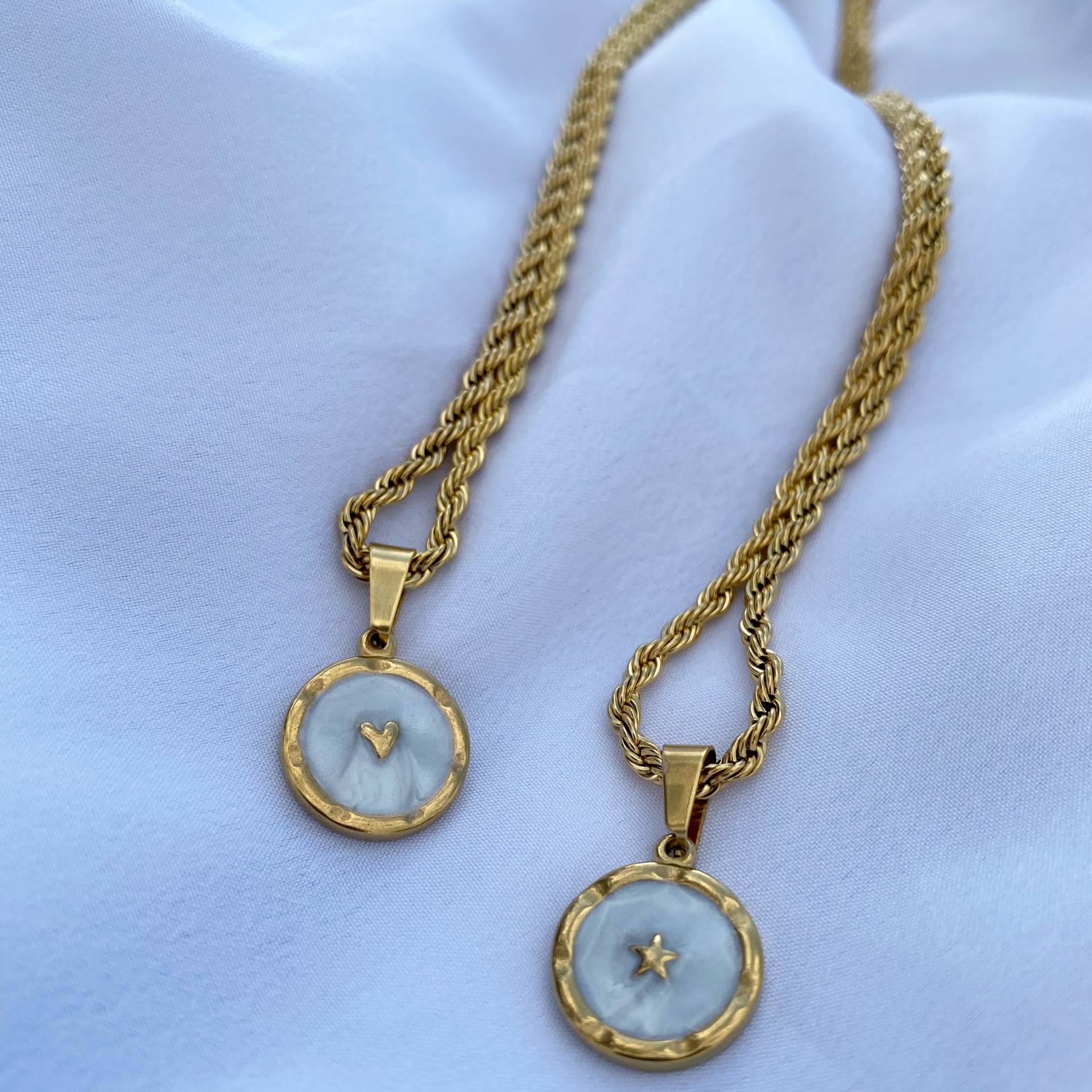 Dois colares dourados com pendentes redondos em madre-pérola com coração e estrela, sobre tecido branco