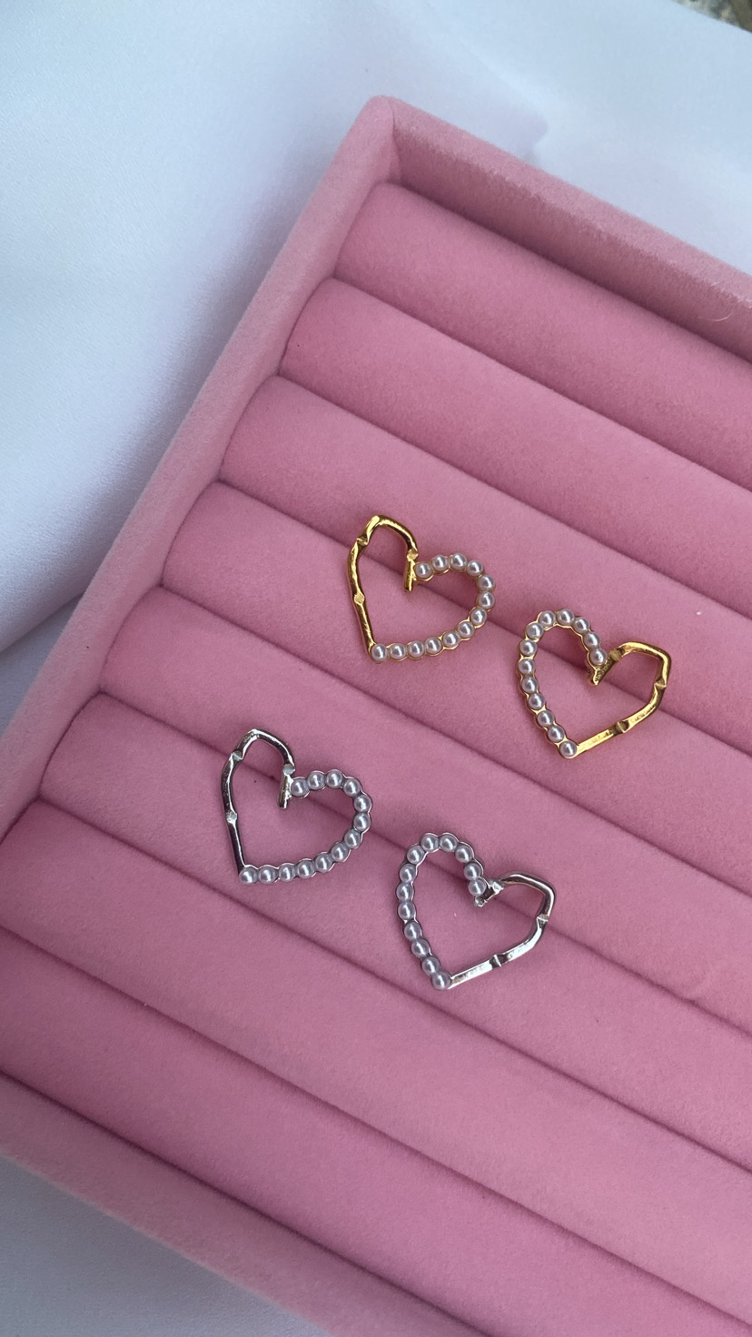 Brincos em forma de coração dourados e prateados com pérolas numa caixa rosa.