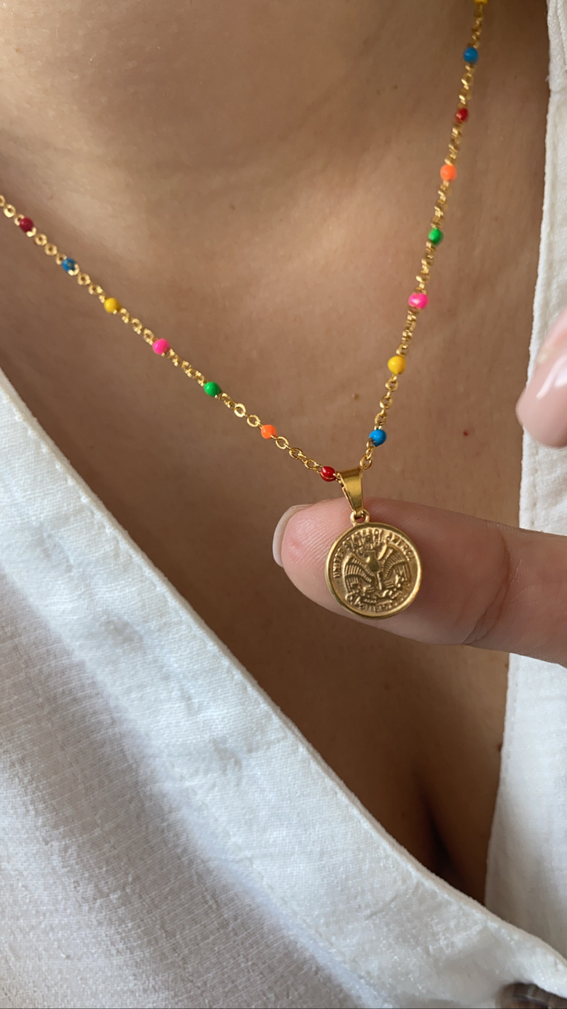 Colar dourado com contas coloridas e pendente redondo em relevo numa pessoa com camisa branca