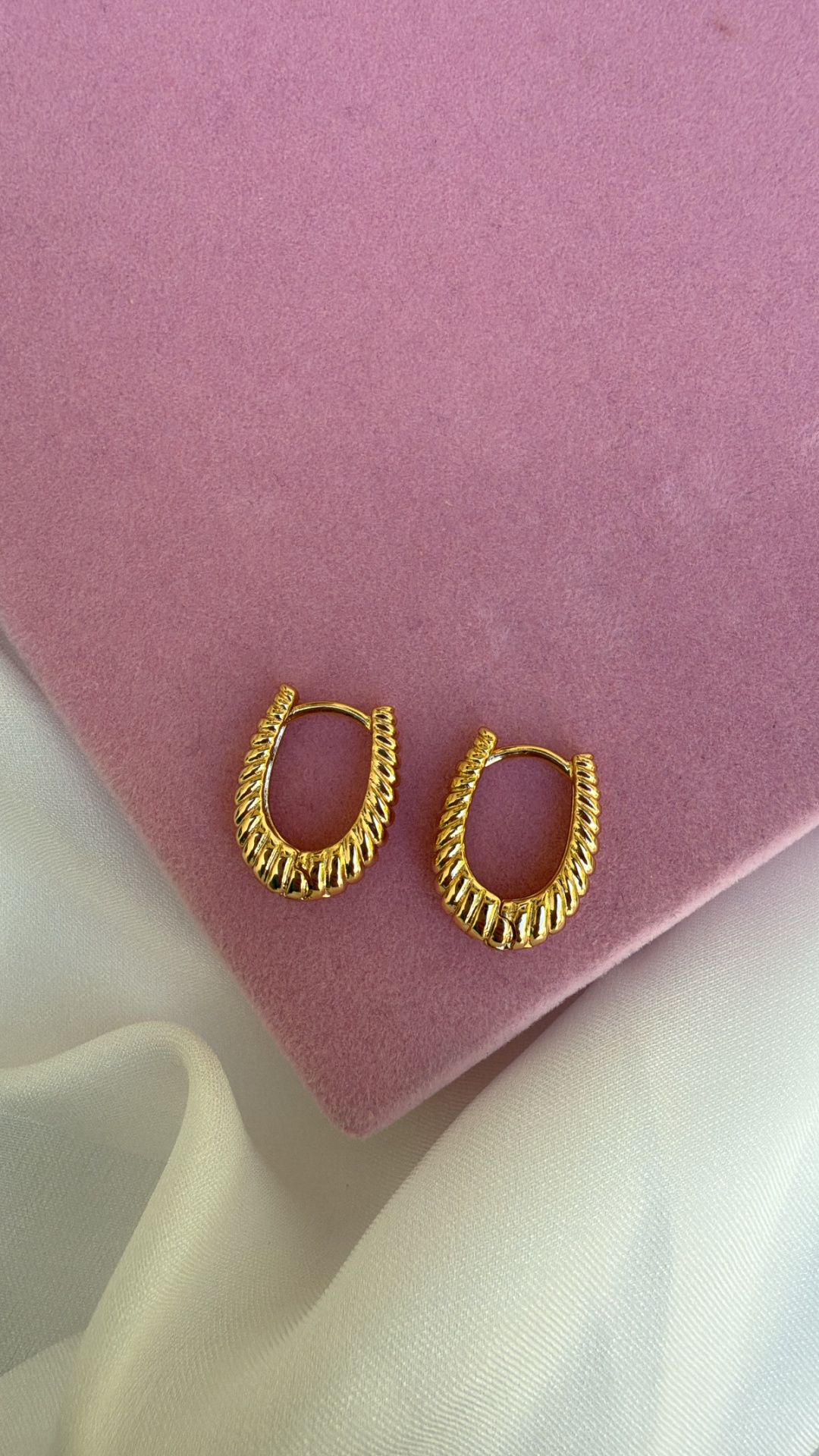 Brincos dourados texturizados em formato oval sobre veludo rosa