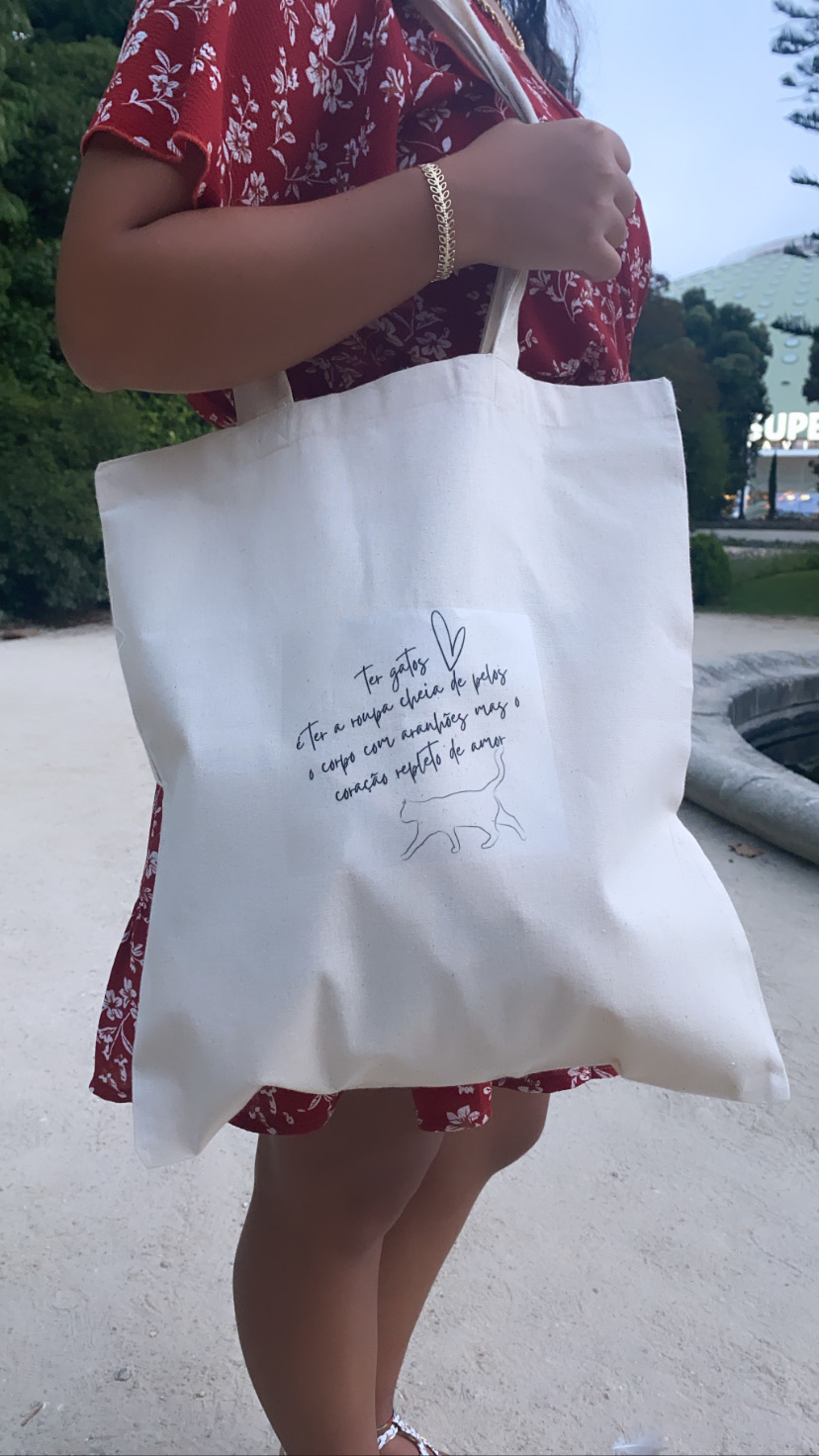 Mala de tecido branco com ilustração e texto sobre gatos, segurada por mulher com vestido vermelho floral, em ambiente exterior