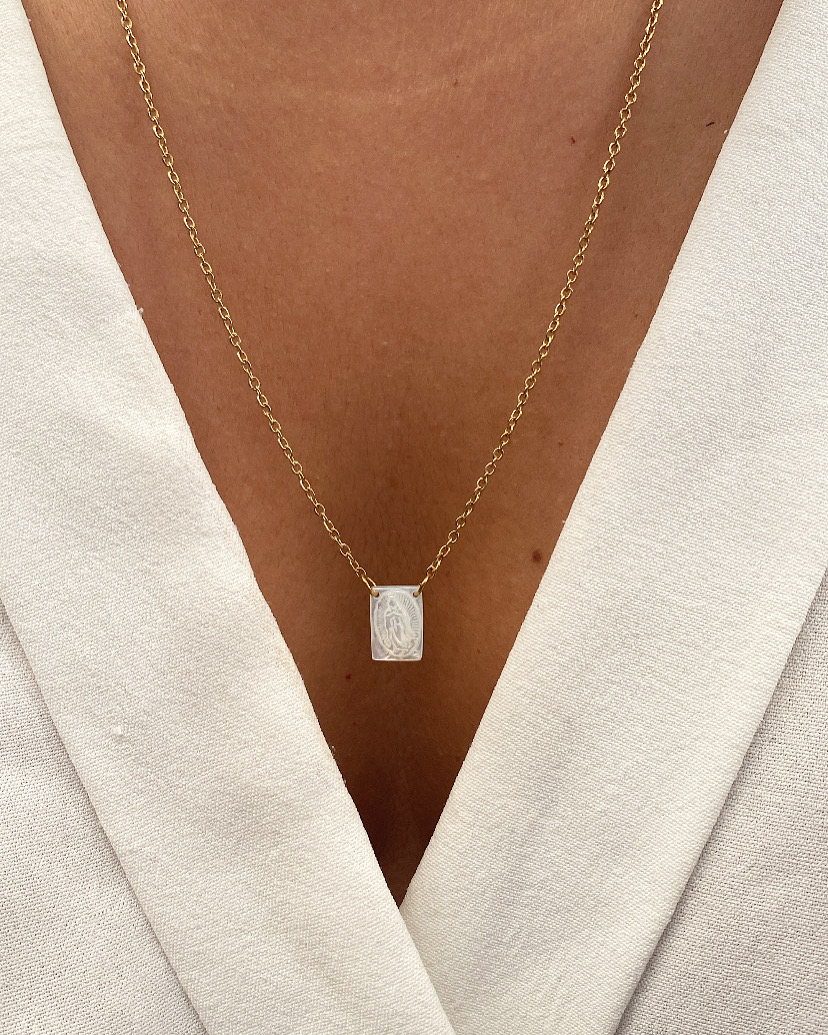 Colar em corrente dourada com pendente retangular prateado no pescoço de pessoa com camisa branca