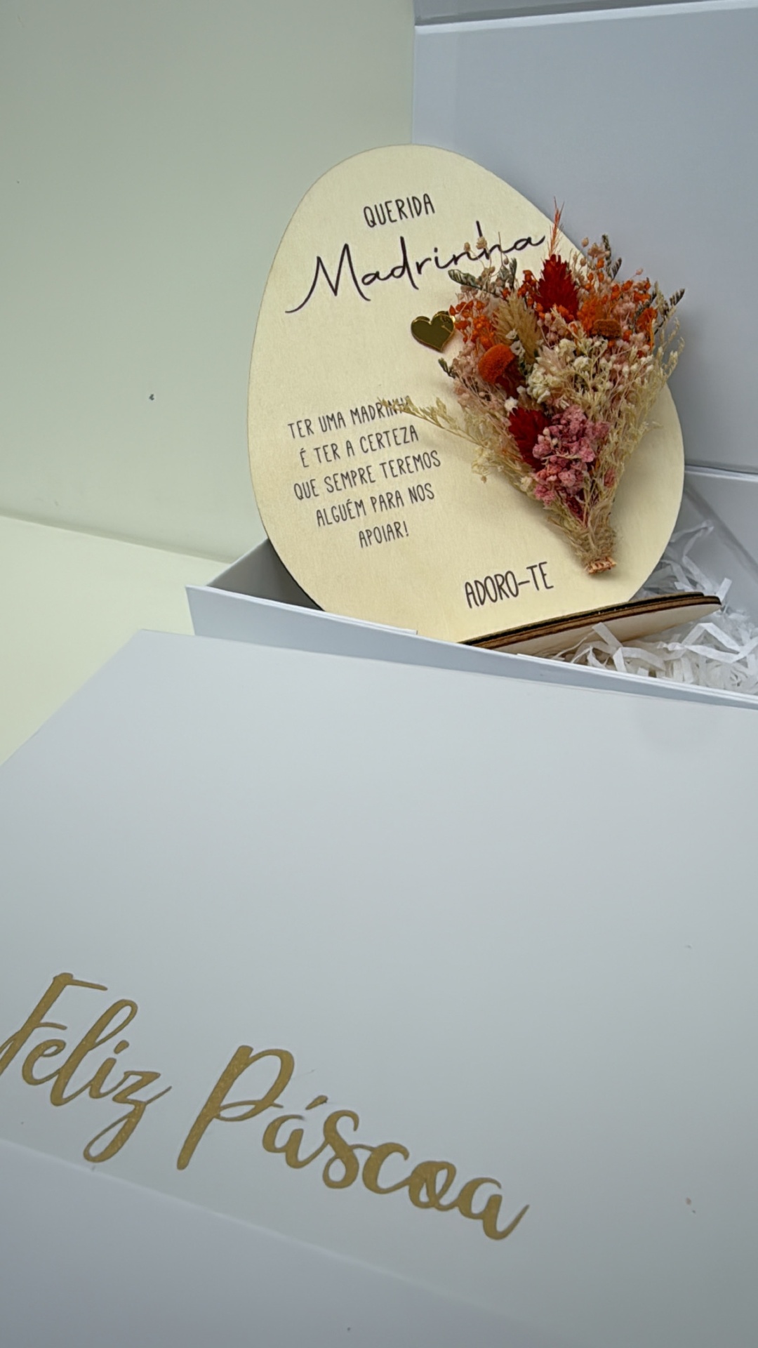 Embalagem branca aberta com decoração de madeira em forma de ovo com flores secas e texto dedicado a madrinha.