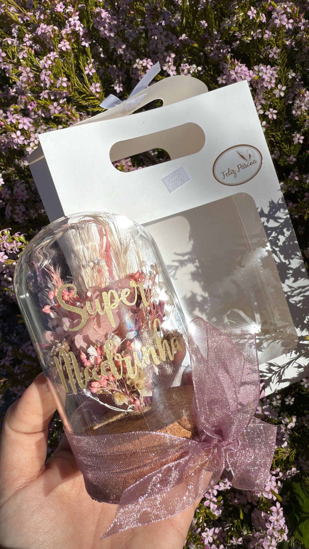 Vaso decorativo de vidro com flores secas e texto 'Super Mãe' em frente a saco branco com texto 'Feliz Páscoa'
