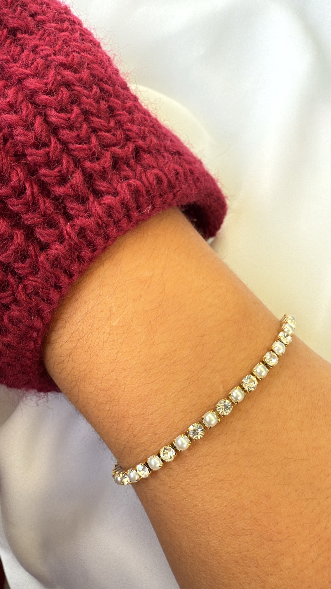 Pulseira tennis com pedras brancas montada em metal dourado no pulso com manga vermelha de malha.