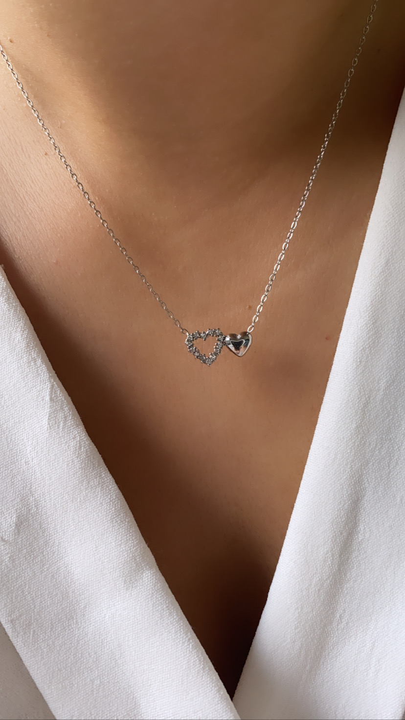 Colar prata com dois corações no pescoço de uma pessoa com camisa branca