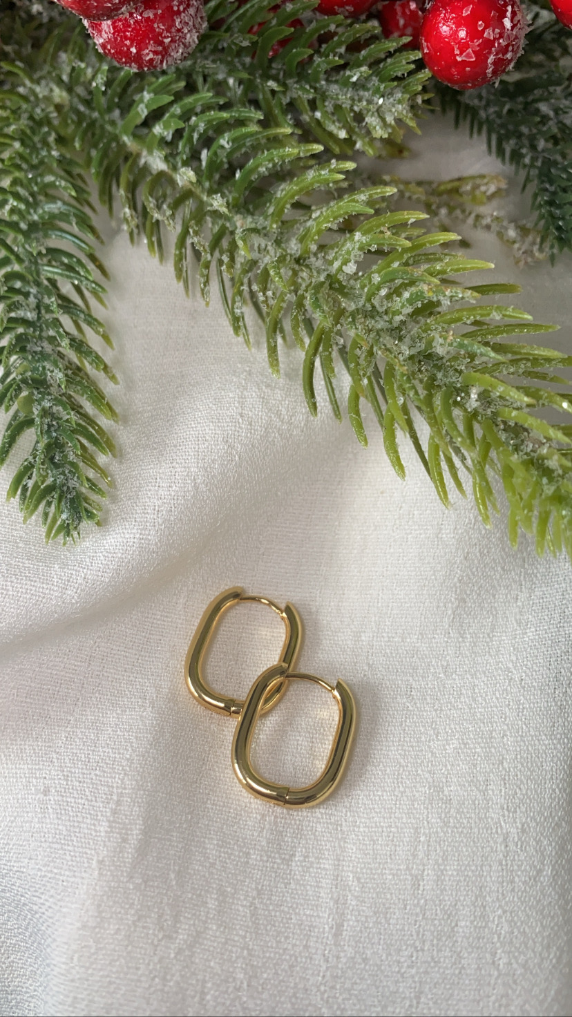 Brincos dourados retangulares sobre tecido branco com ramos de pinheiro e bagas vermelhas