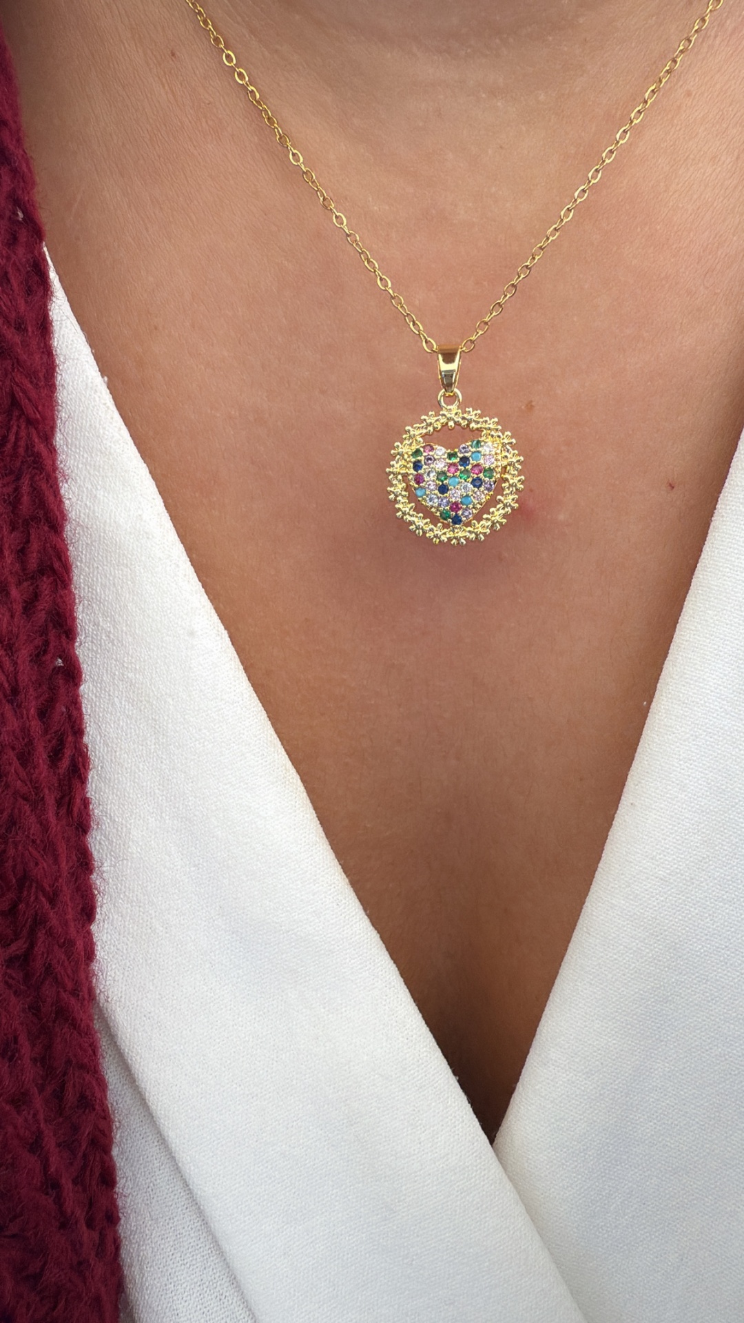 Colar dourado com pendente de coração colorido em fundo branco e cardigan vermelho