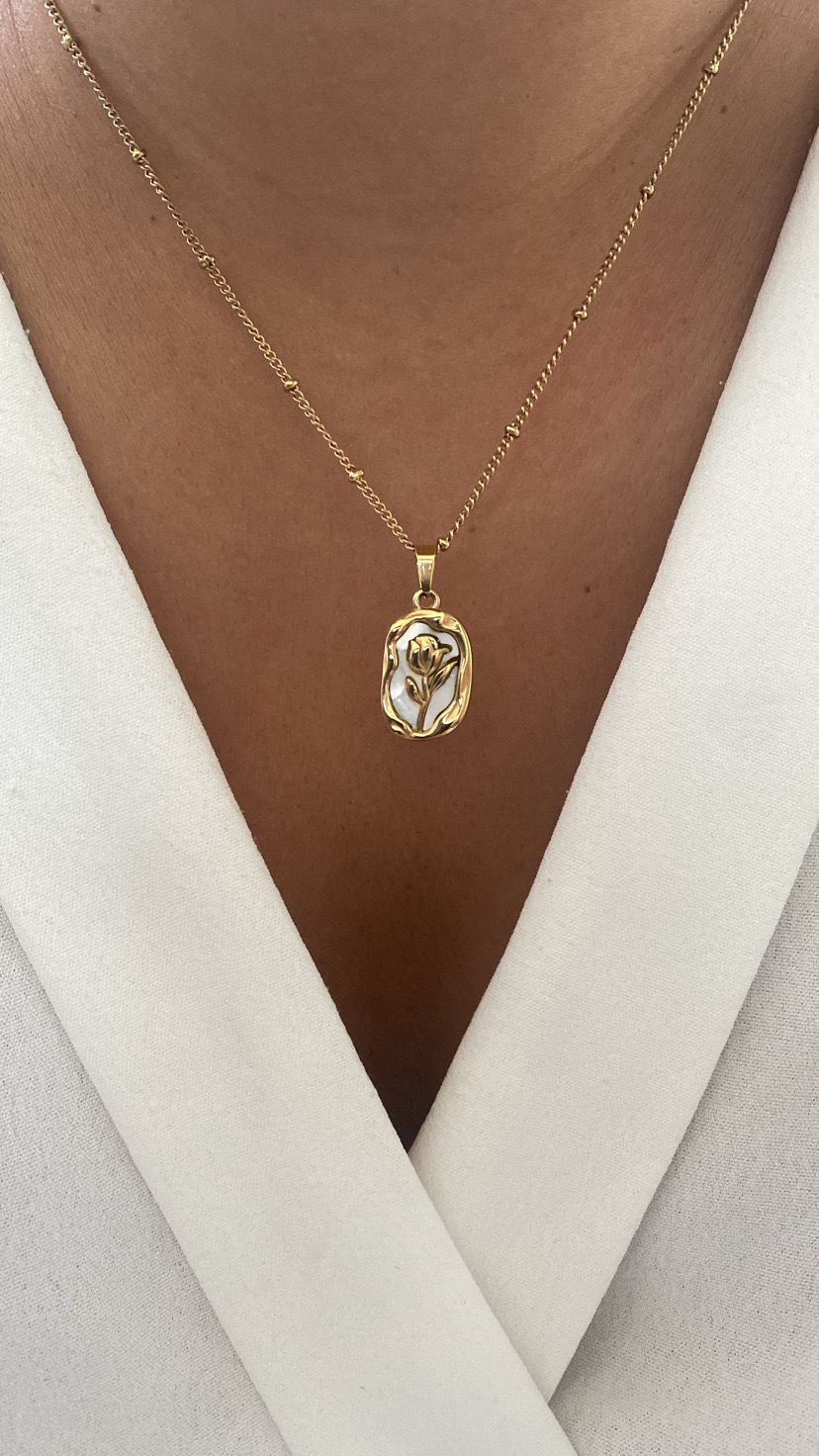 Colar dourado com pendente floral em relevo sobre vestido branco.