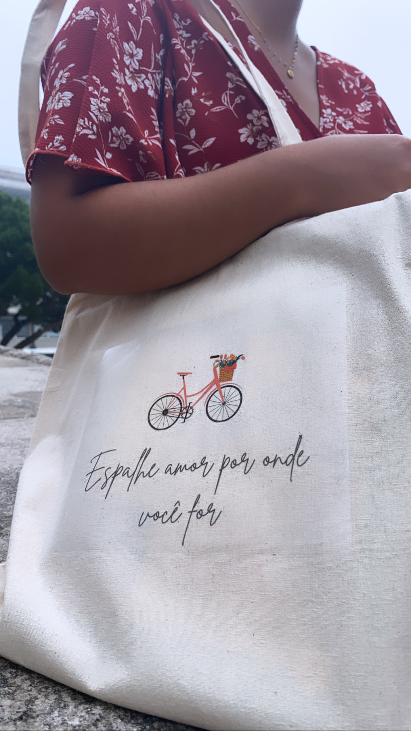 Saco de tecido com estampa de bicicleta e texto, segurado por pessoa de vestido vermelho de flores