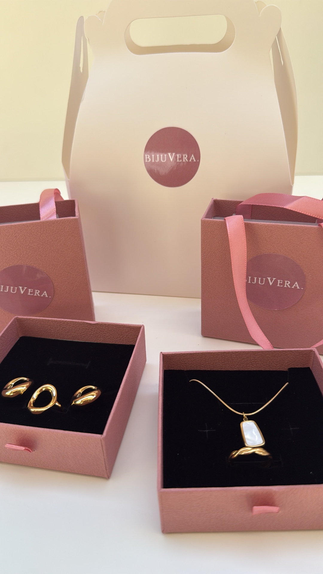 Conjunto de joias douradas com caixas e sacos rosa com logotipo BIJUVERA