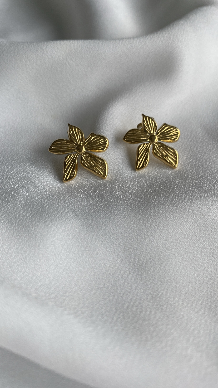 Brincos dourados em forma de flor com quatro pétalas, sobre tecido branco