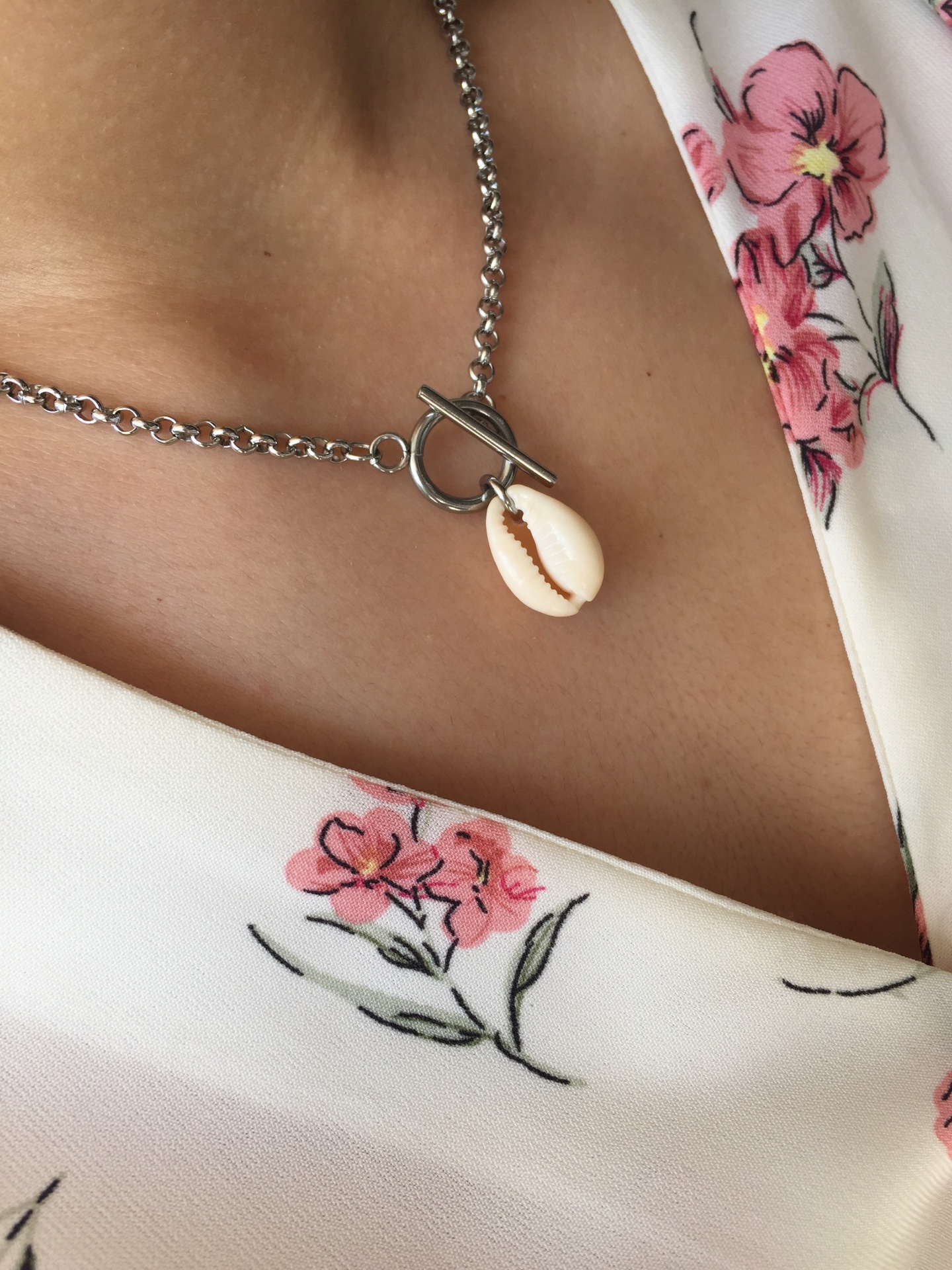 Colar com corrente prateada e pingente de concha sobre roupa branca com flores rosa