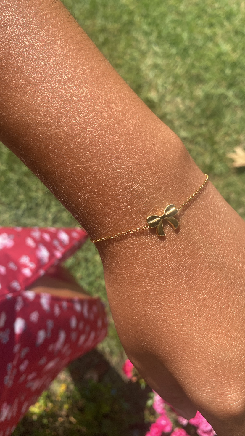Pulseira dourada com pendente em forma de laço no pulso, fundo com relva e roupa vermelha com estampas brancas
