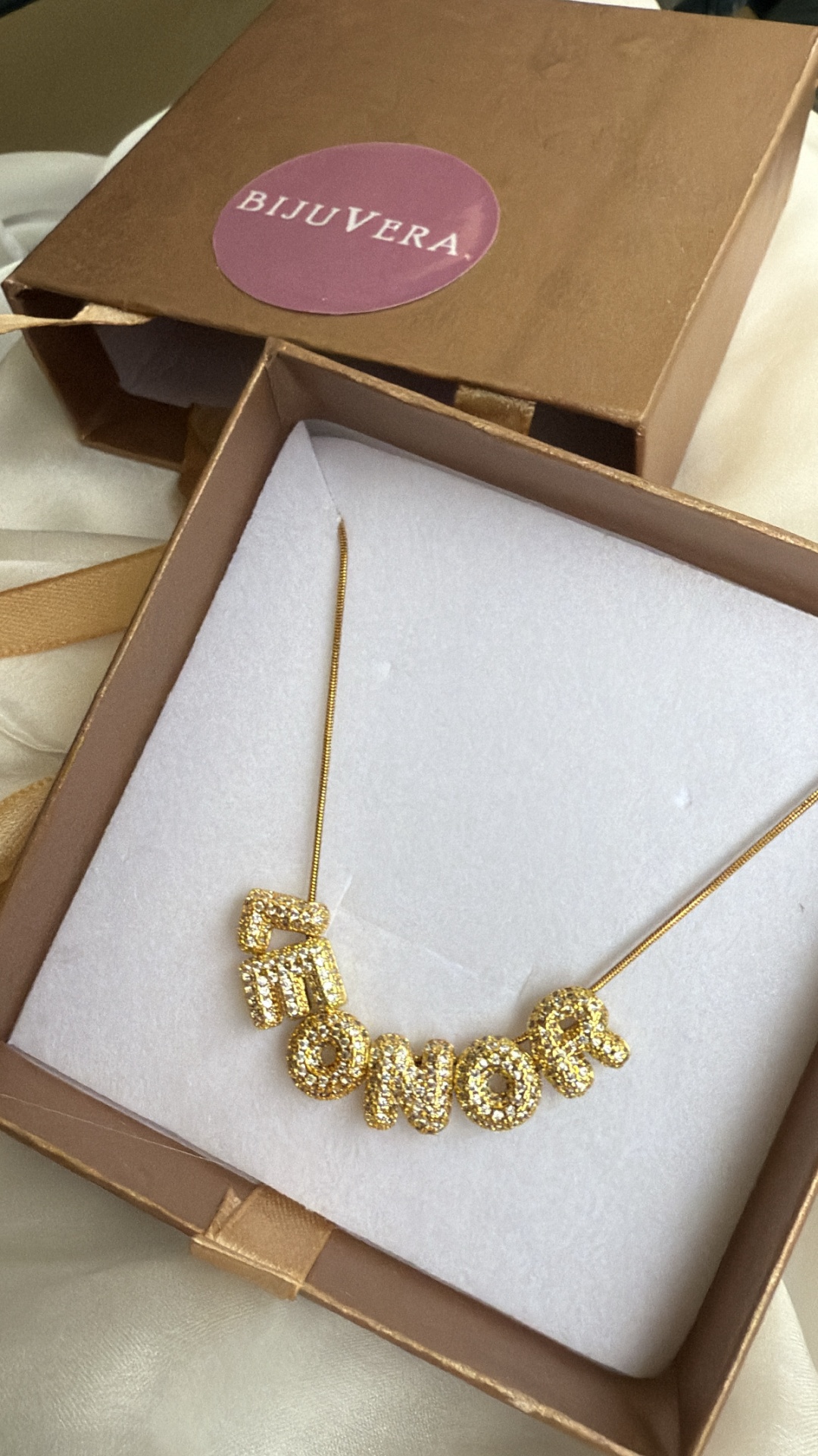Colar dourado com letras cravejadas dentro de caixa castanha com selo BIJUVERA