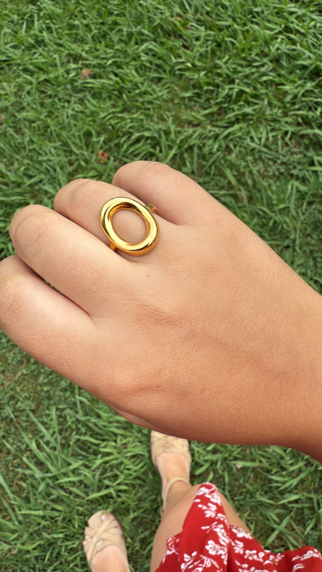 Anel dourado oval numa mão sobre relva verde com vestido vermelho visível
