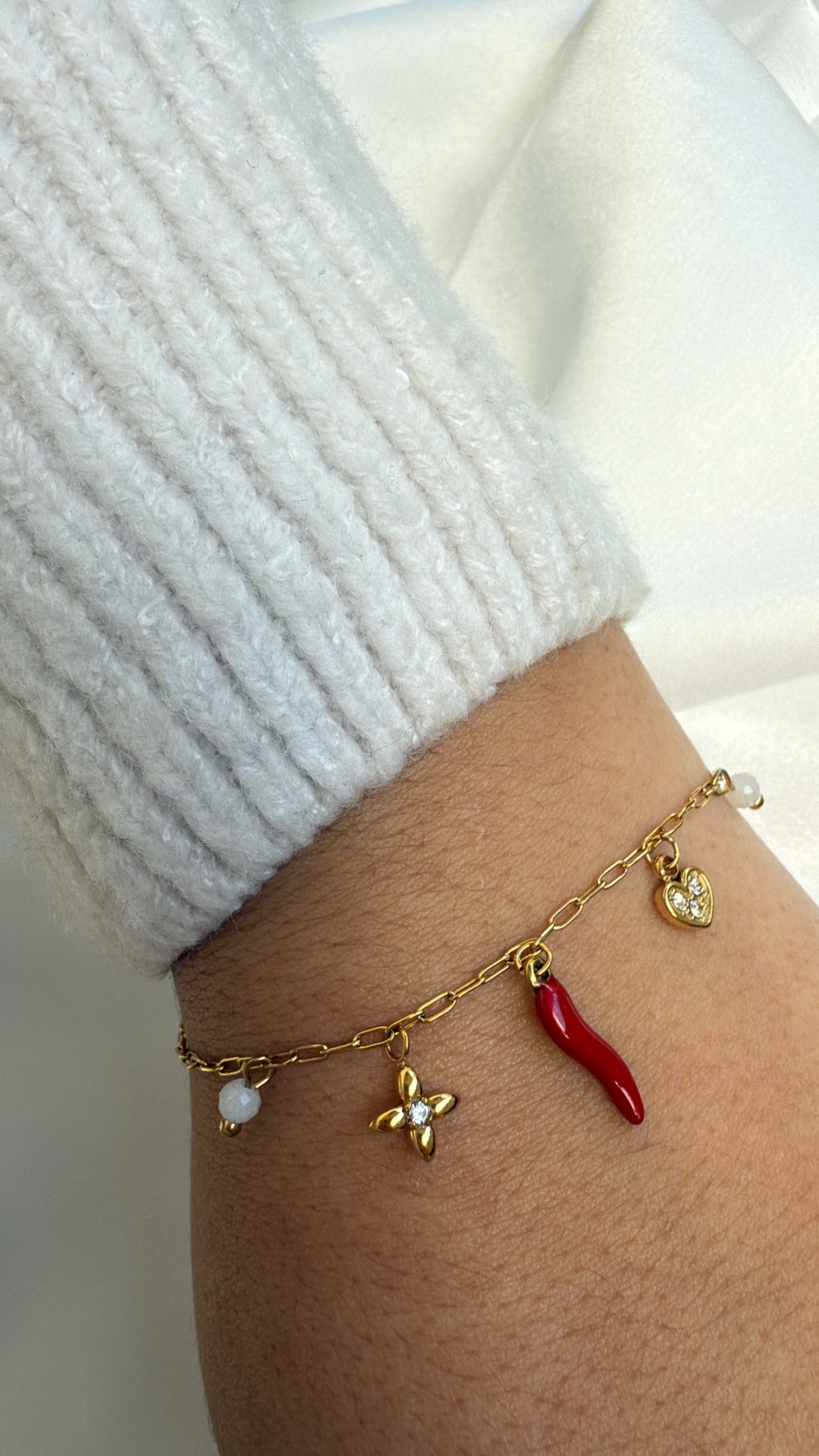 Pulseira dourada com vários pingentes coloridos usada num pulso com manga branca de malha.