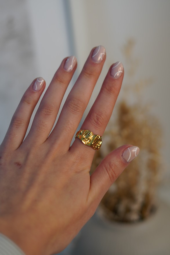 Anel dourado com textura ondulada no dedo anelar de uma mão com unhas pintadas de rosa claro com linhas brancas