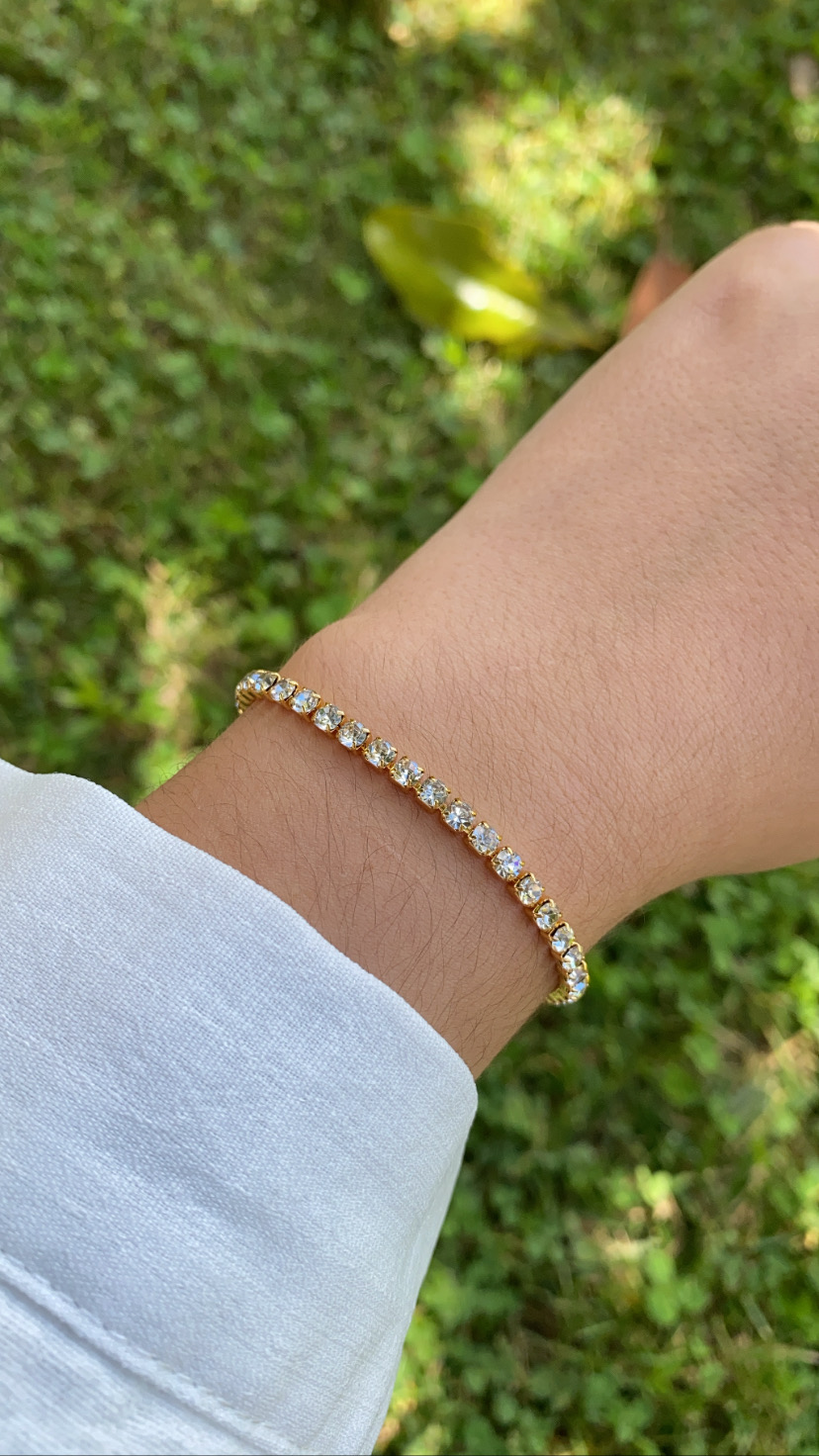 Pulseira tennis dourada com pedras transparentes no pulso em fundo de relva