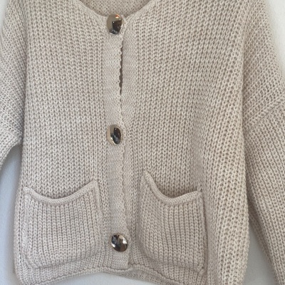 Cardigan bege de malha grossa com botões grandes e bolsos pendurado numa parede branca