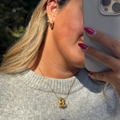 Colar dourado com pendente letra B e brincos dourados em mulher com blusão cinza ao ar livre