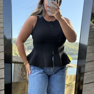 Pessoa a usar top preto de malha com botões e jeans azuis, espelho exterior com paisagem de rio ao fundo