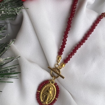 Colar de contas vermelhas com pendente oval dourado em relevo sobre tecido branco com bagas vermelhas e ramos verdes.