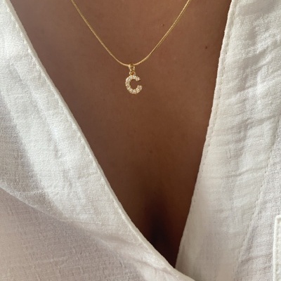 Colar dourado com pendente de letra 'C' em cristais sobre blusa branca