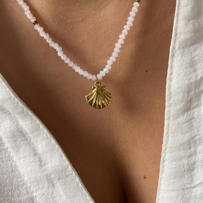 Colar com contas brancas e douradas e pendente dourado em forma de concha sobre blusa branca.