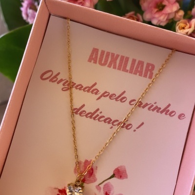 Colar dourado com pendente em pedra transparente em caixa rosa com mensagem de agradecimento e flores ao fundo