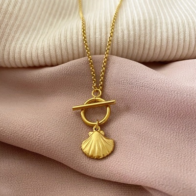 Colar dourado com pendente de concha sobre tecidos creme e rosa.