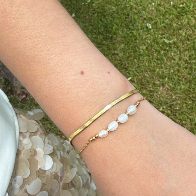 Pulseira dourada lisa e pulseira com contas brancas no pulso num outdoor com relva