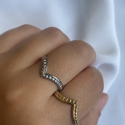 Dois anéis em V, um prateado com pedras e outro dourado texturizado, na mão sobre tecido branco