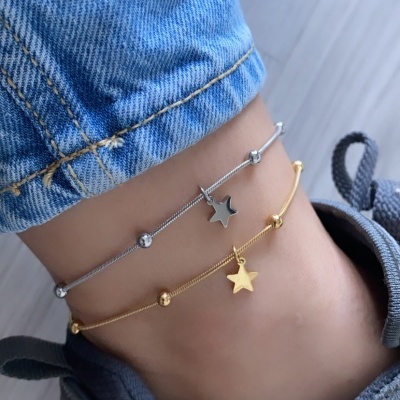 Tornozelo com duas pulseiras finas, uma prateada e outra dourada com pendentes de estrelas, calça de ganga azul e sapatilha cinzenta